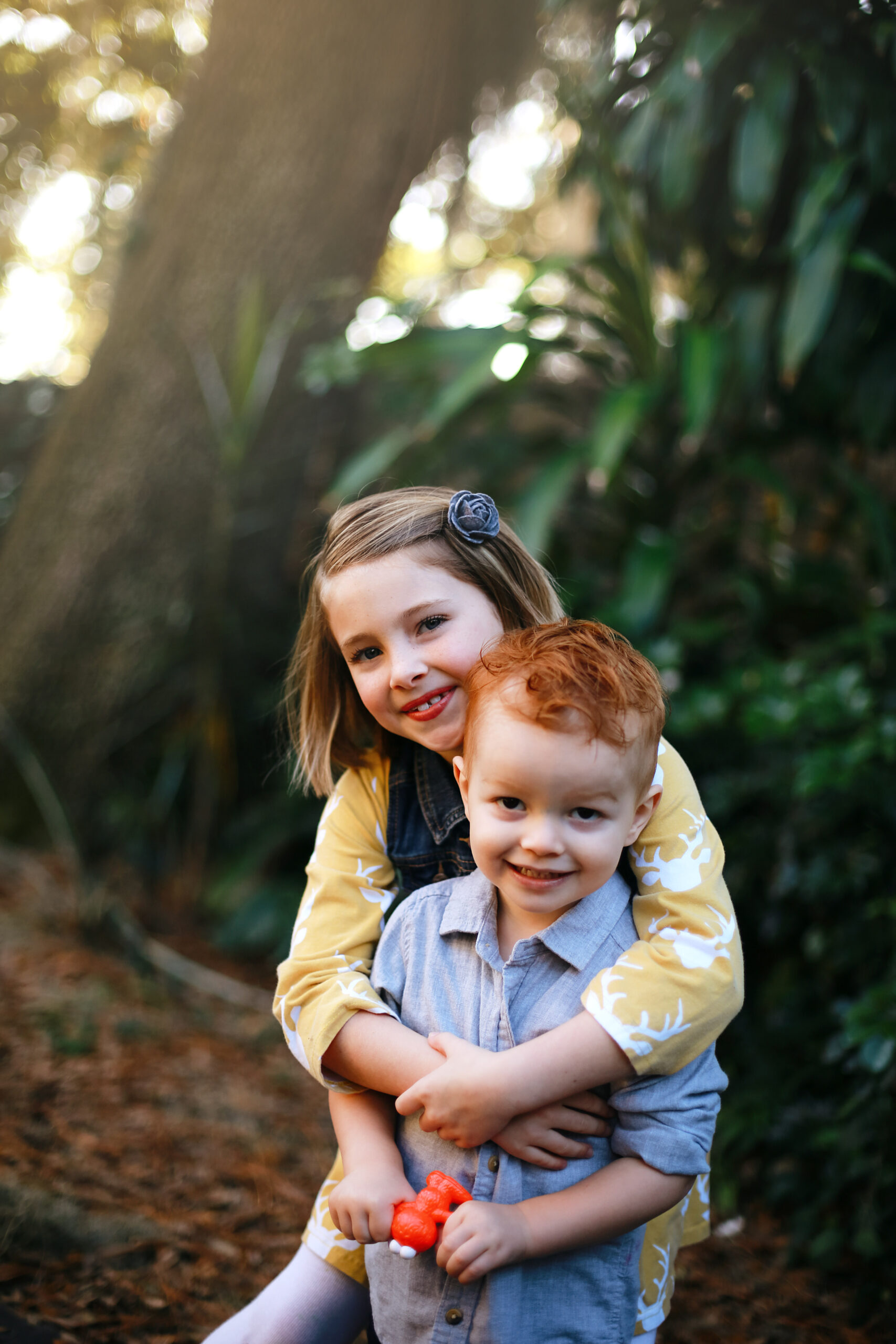 two kids standing outside, one young blonde girl and one redheaded toddler boy and the girl is hugging the boy from behind and they're both smiling at the camera