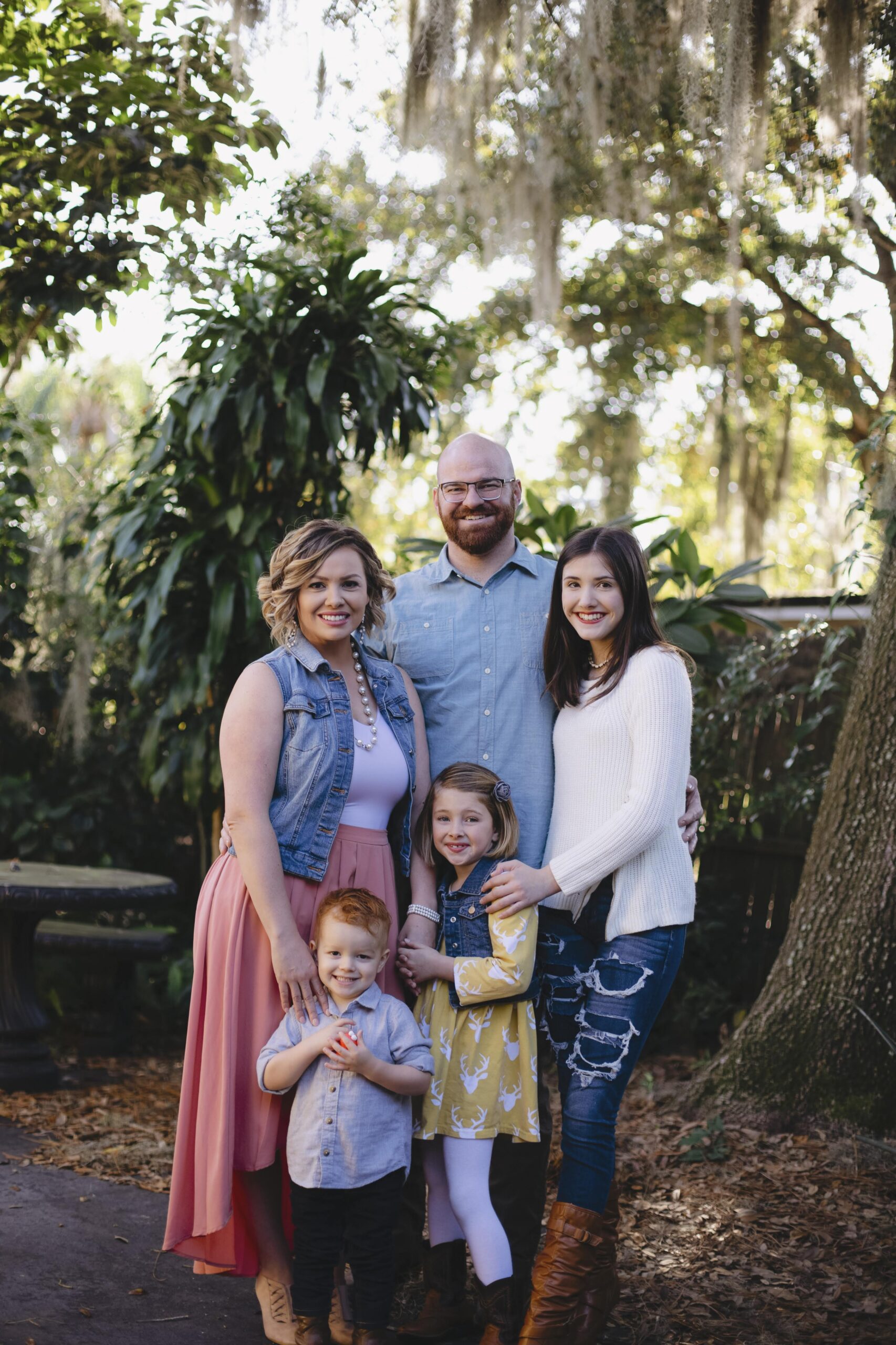 family photo (taken outside) of 5 family members. a blonde & brown curly haired mom, a bald dad with a beard, a brunette teenage girl, a blonde young girl and a redheaded toddler boy.