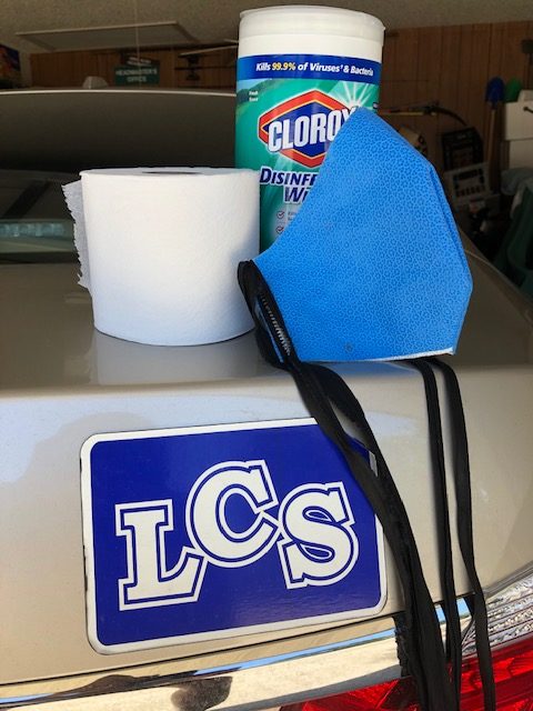 clorox wipes, toilet paper and a face mask sitting on the trunk of a car with an LCS sticker/magnet on the back of the car