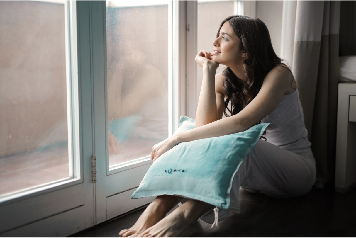 woman sitting next to a window looking out the window, holding a pillow and wearing relaxed clothes and no shoes