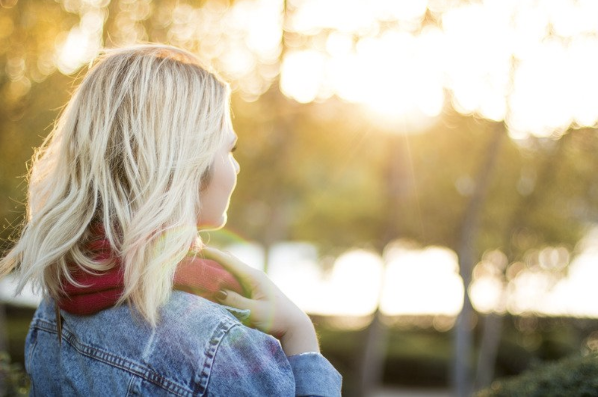 picture of blonde woman at sunrise or sunset looking into nature surrounded by trees wearing a red scarf and a jean jacket 