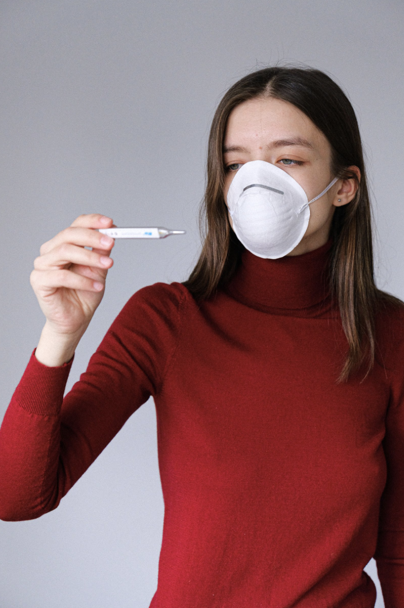 brunette woman wearing a red turtleneck, long sleeved shirt with a face mask looking at a thermometer 