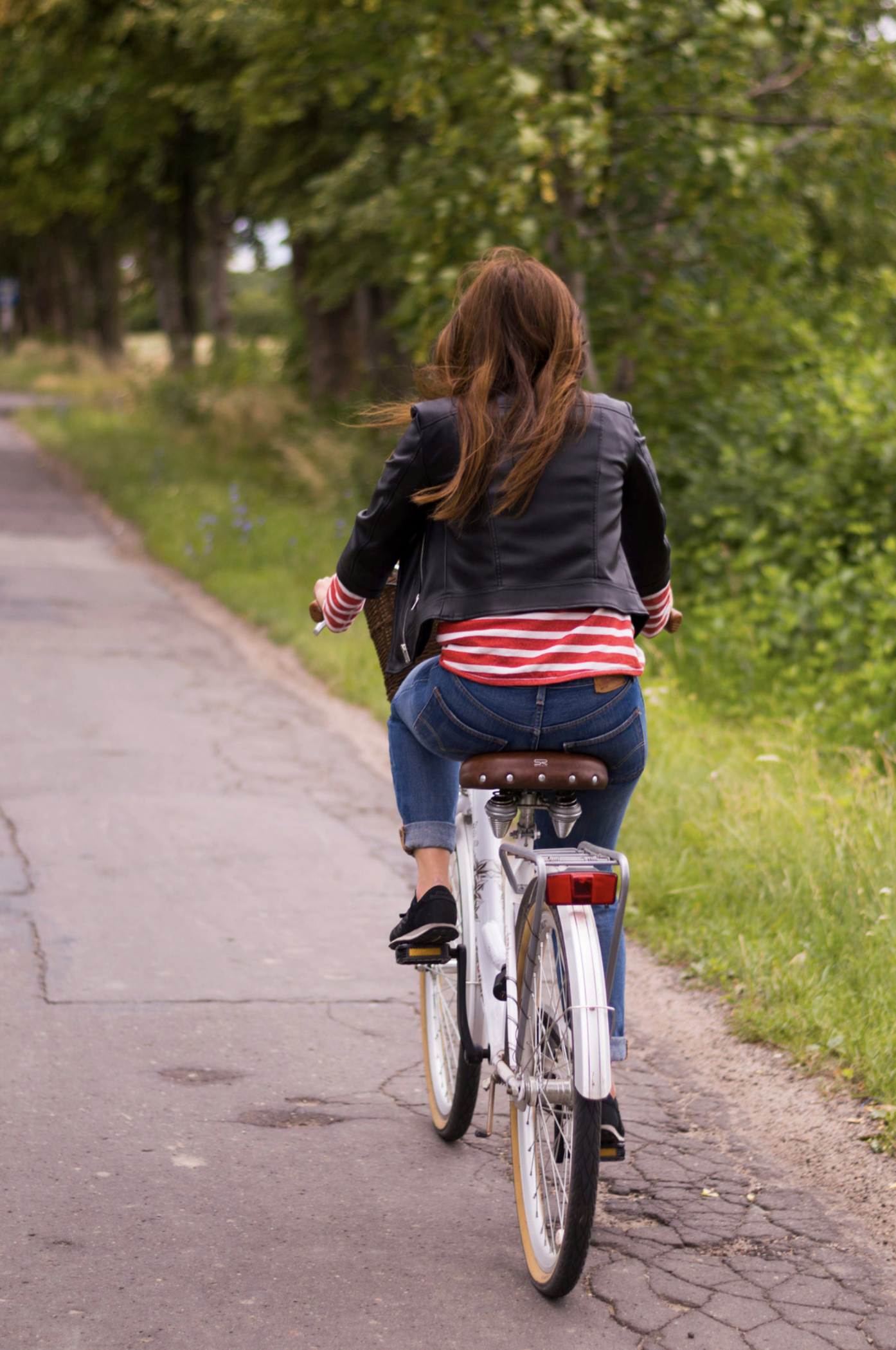 rear view of a young woman riding a bike on an old road with green grass and trees surrounding the road. She has long brunette hair blowing in the wind and is wearing a black leather jacket with a white and red stripped shirt, jeans and black shoes