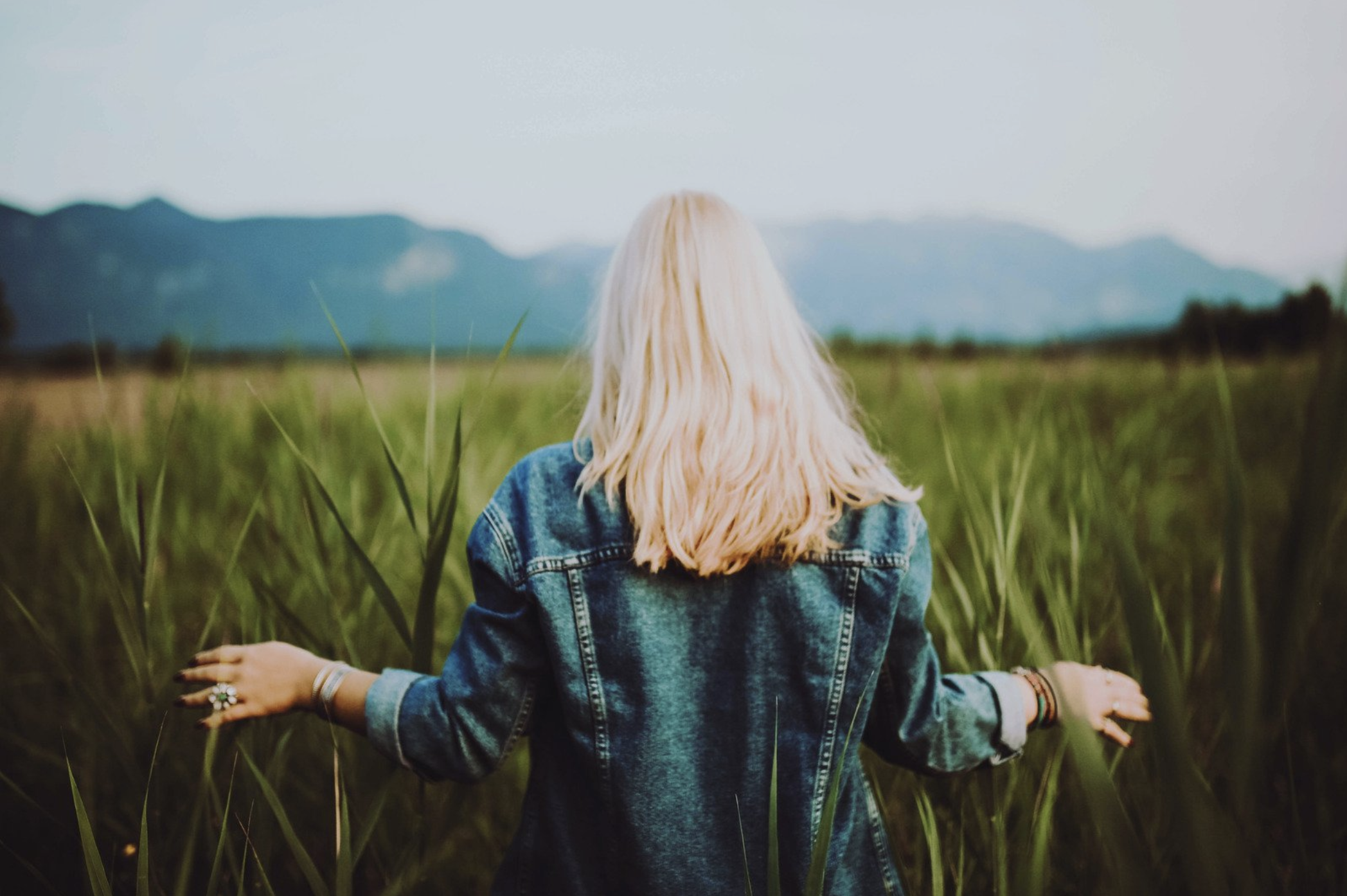 Blonde young woman walking through a field of tall green grass with her hands out feeling the grass and feeling the wind blow her hair