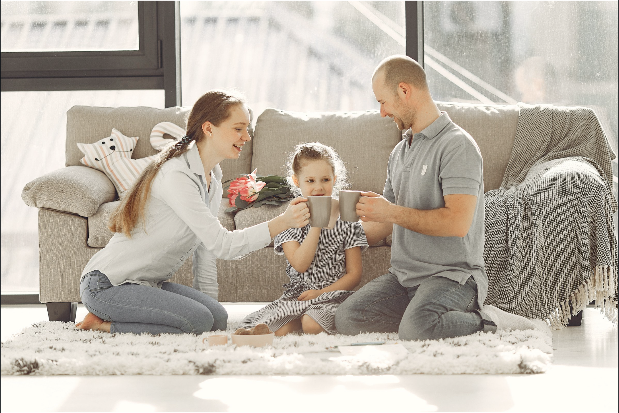 three people sitting on the floor in front of a couch with coffee cups. A mom, a daughter and a dad all looking at each other smiling.